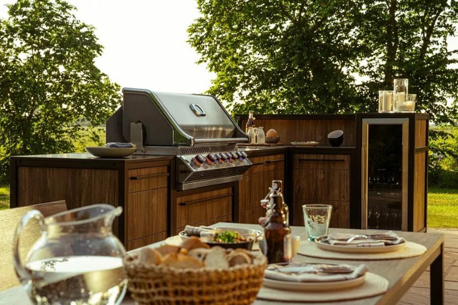 Outdoorküche aus dunklem Holz mit integriertem Gasgrill und Kühlschrank. Im Vordergrund steht ein gedeckter Tisch und im Hintergrund Bäume und Felder.