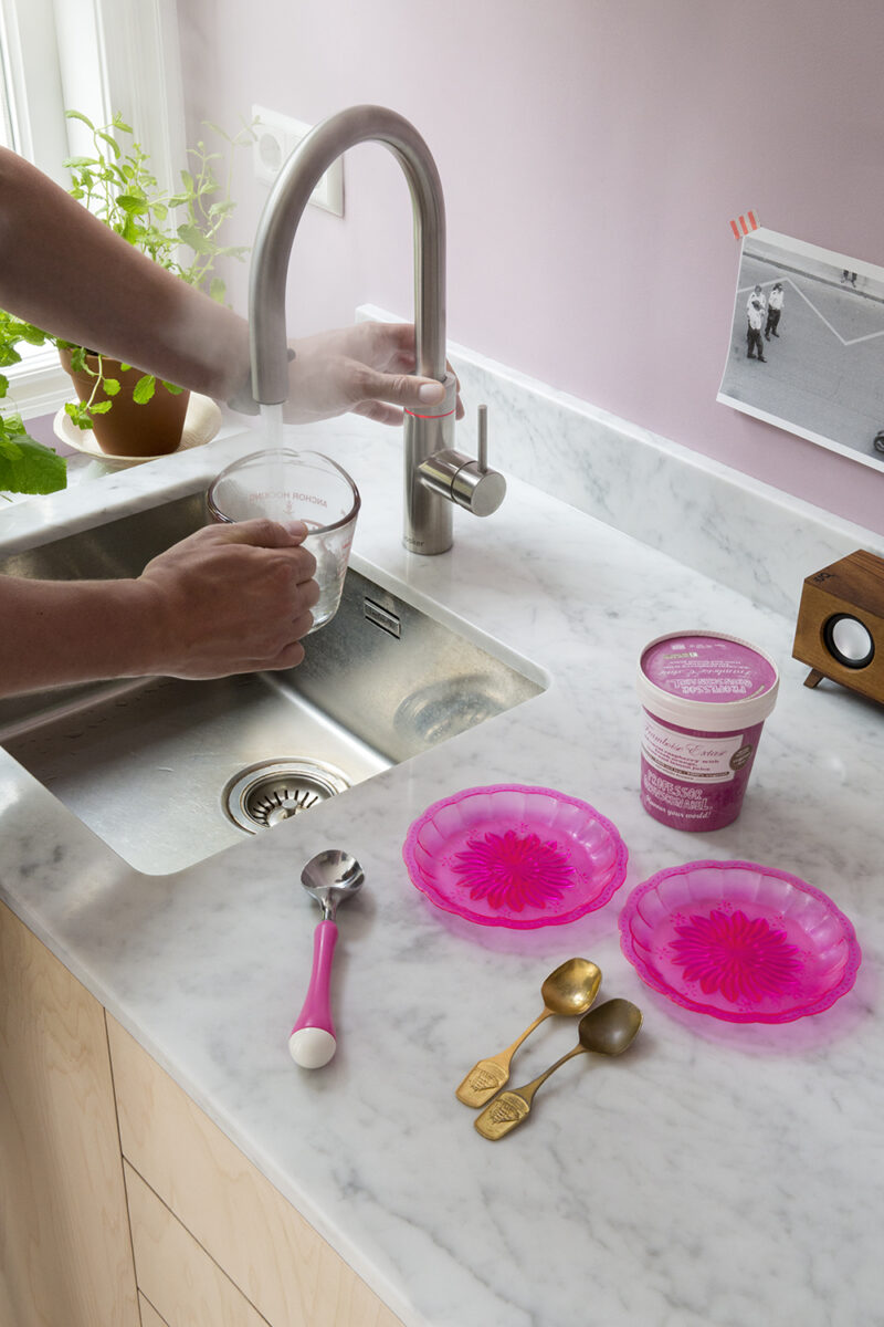 Ein Quooker-Wasserhahn in Edelstahloptik wird verwendet, um heißes Wasser in einen Messbecher zu füllen. Auf der Arbeitsplatte liegen Eisbecher und Löffel.