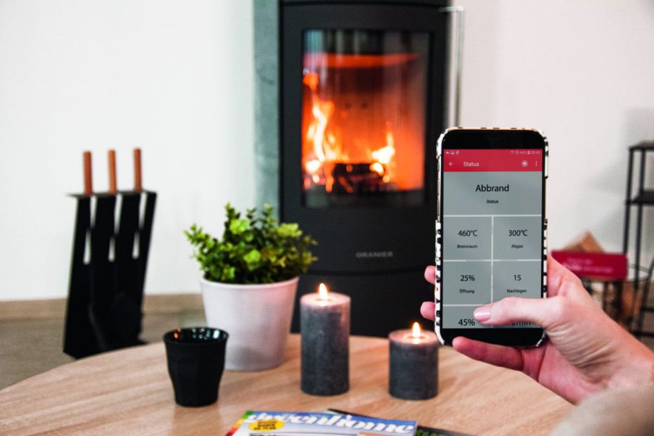 Smartphone zeigt App-Anzeige zur Abbrandsteuerung conFlame, im Hintergrund ORANIER Kaminofen im Betrieb mit sichtbarem Feuer.