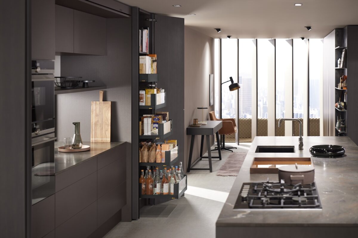 Moderne, offen gestaltete Küche mit dunklen Fronten und einer großen Kücheninsel; ein hoher Vorratsschrank mit mehreren herausgezogenen Regalböden ist geöffnet und zeigt Lebensmittel. Im Hintergrund befindet sich ein heller Wohnbereich mit Schreibtisch, Sessel und bodentiefen Fenstern.