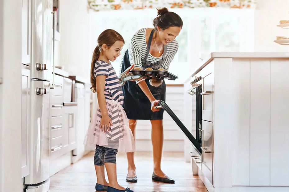 Ein helles, warmes Lifestyle-Bild für das Beitrags-Header. Es zeigt eine Frau in einer dunkelgrauen Schürze und ein Mädchen in einem gestreiften Kleid, die Muffins in einer modernen, weißen Küche backen. Der Backofen steht offen. Das Bild symbolisiert die Freude am Kochen und die Alltagstauglichkeit moderner Küchengeräte.