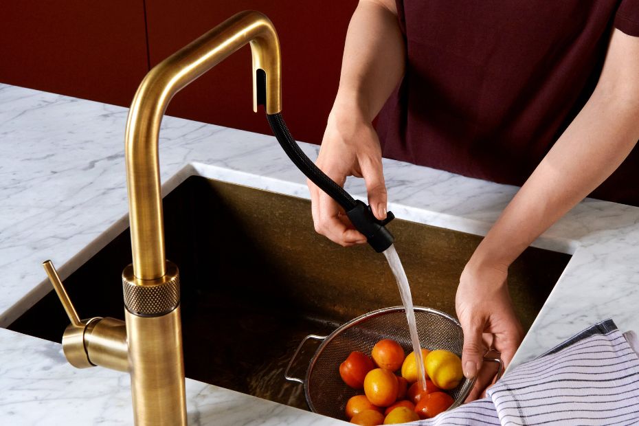 Messingfarbene Küchenarmatur an einer Spüle, während Obst unter fließendem Wasser gewaschen wird