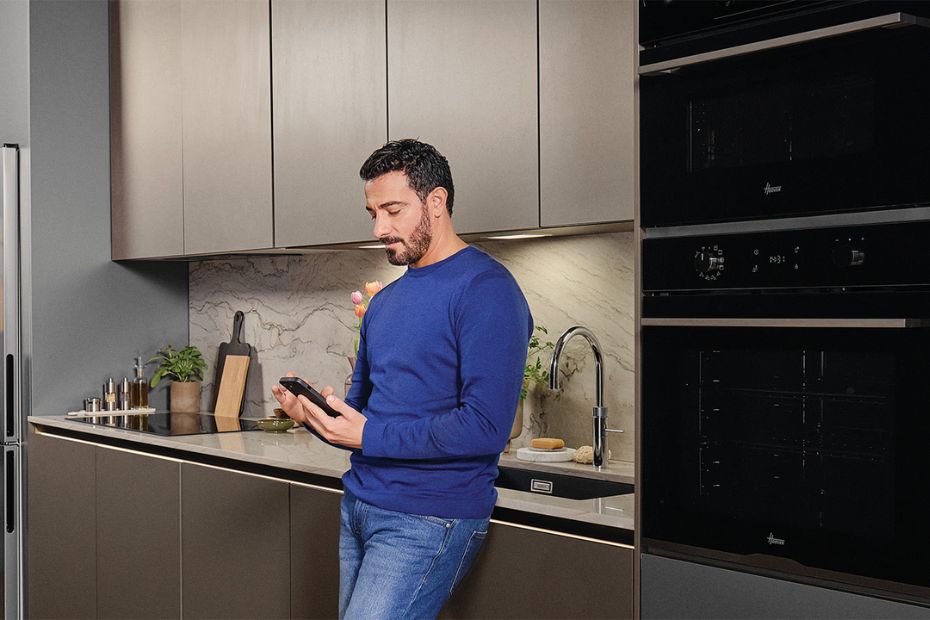 Person mit Smartphone in moderner Küche mit Hoover Einbaugeräten, Backofen und Kühlschrank
