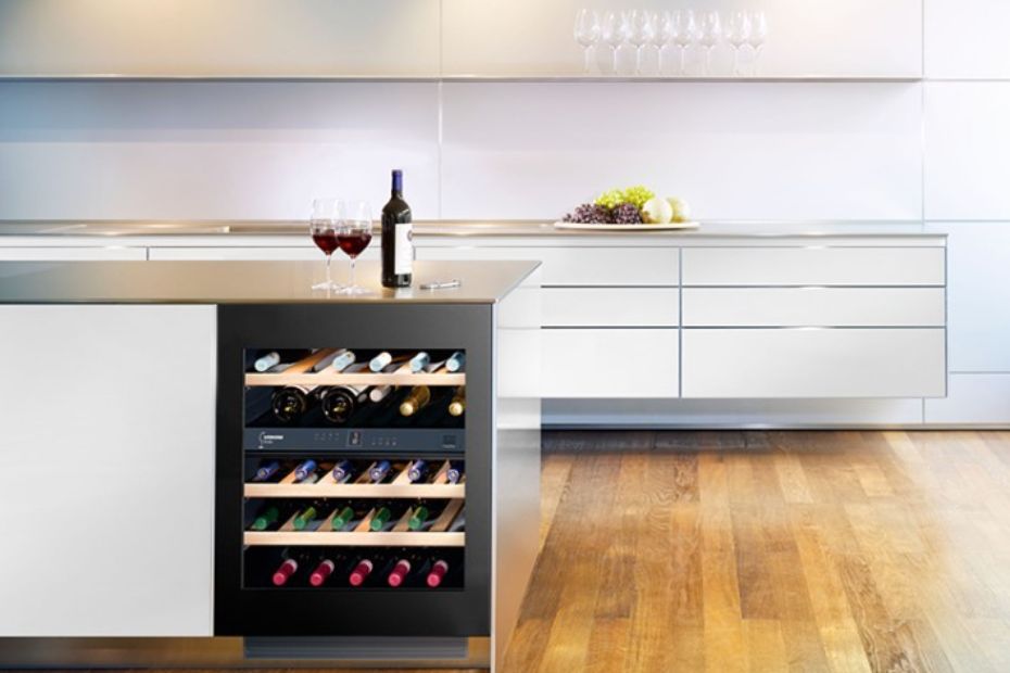 Unterbau-Weinkühlschrank mit Glastür und Lüftungsgitter unter der Tür in einer weißen Küche; darüber stehen eine Weinflasche und zwei Gläser.